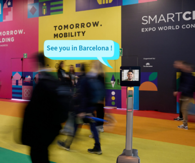 Digital montage showing a 5G Metro telepresence robot added to a photo of the Smart City Expo World Congress venue in Barcelona. Blurred visitors walk through the colourful exhibition space, while the robot displays a student on its screen and a speech bubble reading “See you in Barcelona!”.