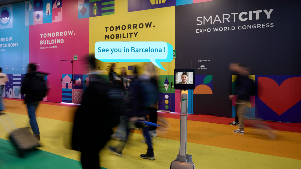 Digital montage showing a 5G Metro telepresence robot added to a photo of the Smart City Expo World Congress venue in Barcelona. Blurred visitors walk through the colourful exhibition space, while the robot displays a student on its screen and a speech bubble reading “See you in Barcelona!”.